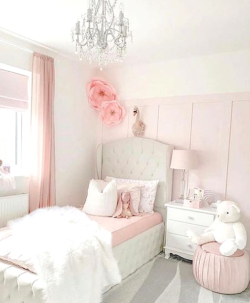 a light pink and white girl's bedroom with a white upholstered bed with pink and bedding, a white nightstand, a pink pouf and a crystal chandelier