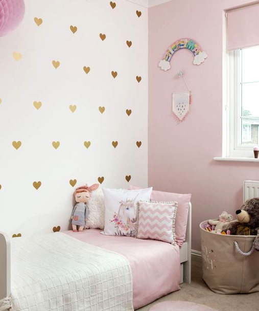 a pretty girl's bedroom with a light pink wall, a gold heart accent wall, a white bed with pink and white bedding, a basket with toys