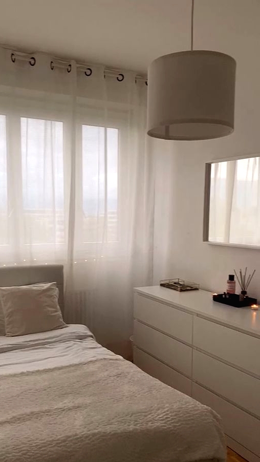 a small and cool minimalist bedroom done in neutrals, with a sleek white dresser, a pendant lamp, a creamy bed with neutral bedding, a mirror in a white frame