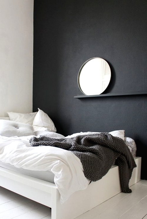 a minimalist bedroom with a black accent wall, a white bed with monochromatic bedding, a shelf with a round mirror and nothing else
