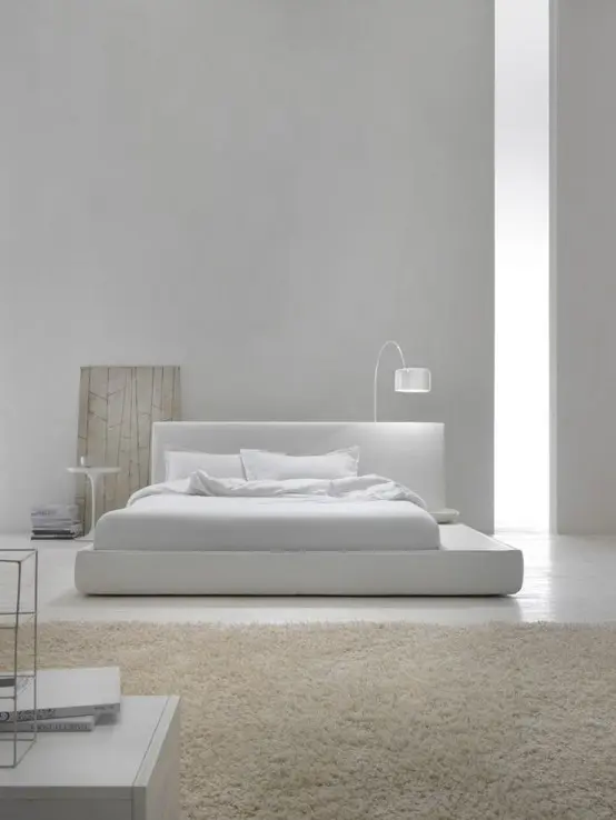 a neutral minimalist bedroom with a sleek bed with neutral bedding, a tan rug and sleek white furniture plus a small white lamp