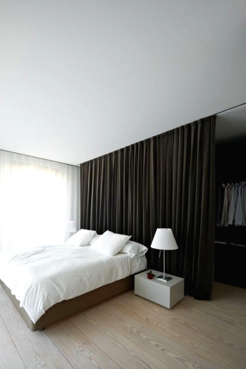 a minimalist bedroom with a walk-in closet hidden with a large dark curtain and a neutral bed with white bedding, a sleek nightstand with a white lamp and much natural light