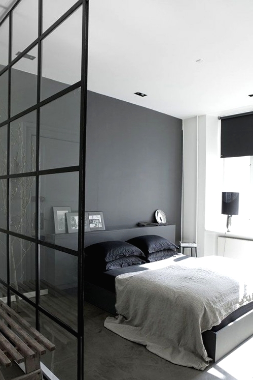a Scandi meets minimalist bedroom with a grey accent wall, a grey bed with a storage headboard, black bedding and black lamps plus a glass space divider