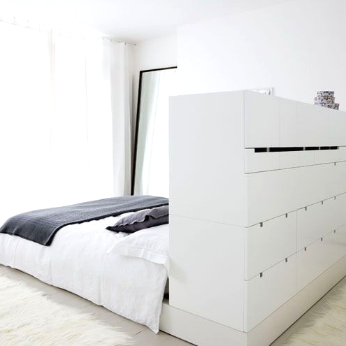 a minimalist white bedroom with a bed with a storage headboard, with lots of drawers, a floor mirror and a glazed wall with curtains