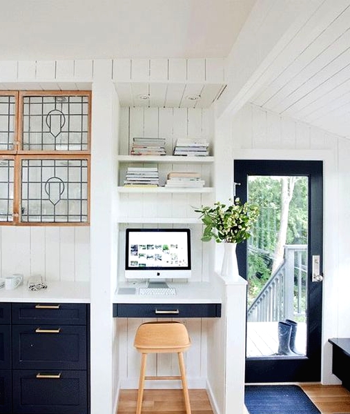 a farmhouse kitchen with navy cabinets, a small nook with a built-in desk, built-in shelves, a light-stained stool and potted greenery