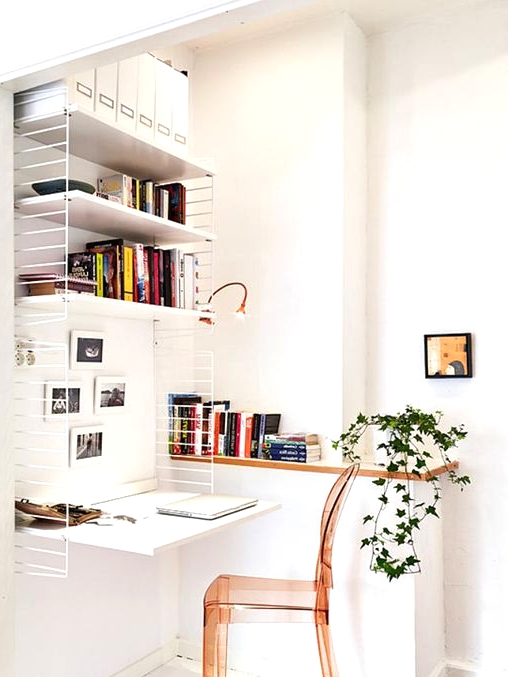 a small and cool nook turned into a home office with a large shelving unit with lots of books, a desk here, an amber acrylic chair and greenery