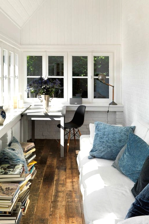 a Nordic sunroom and a home office in one, with a windowsill desk, a white sofa with blue pillows, lots of books and some blooms