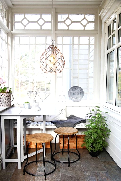 a Nordic sunroom in white, with grey tiles on the floor, a built-in bench, a white table, wooden stools and a woven pendant lamp