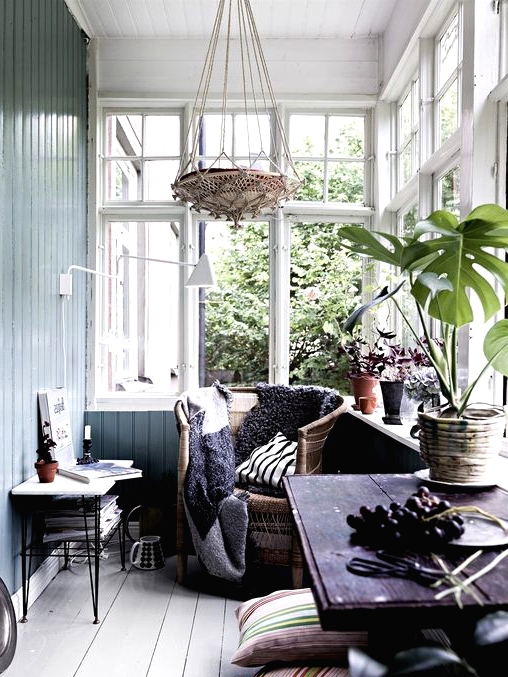 a Nordic sunroom with light blue walls, a dark stained table, a rattan chair, a woven pendant lamp, printed pillows and potted plants