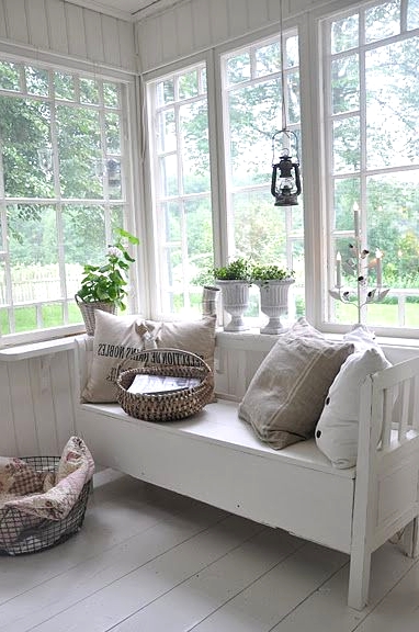 a white Nordic sunroom with a vintage bench, neutral pillows, a wire basket, potted greenery, a pendant lamp