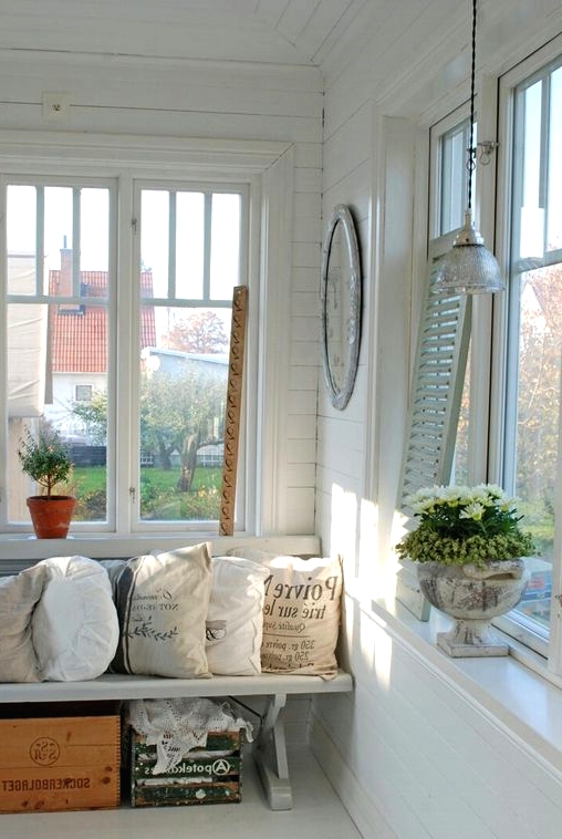 an eclectic Scandinavian sunroom done in white, with planked walls and a floor, a white bench, printed pillows and crates for storage, shutters and a pendant lamp