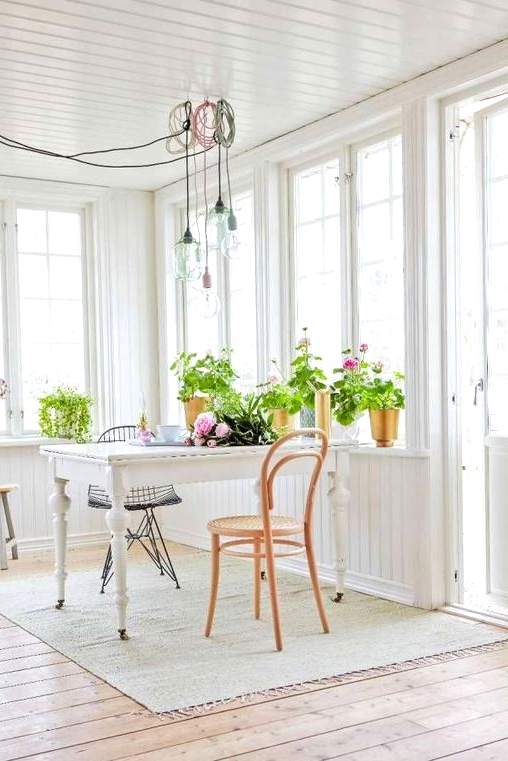an airy white Scandinavian sunroom with a vintage dining table, vintage chairs, pendant lamps, potted plants and blooms