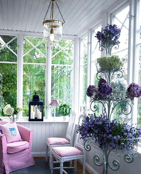 a vintage Scandinavian sunroom with white planks, a printed chair and stools, a vintage metal stand with lots of blooms