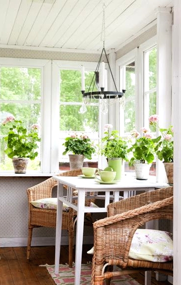 a Nordic sunroom with a white table and wicker chairs, potted greenery and blooms and a printed plant is amazing
