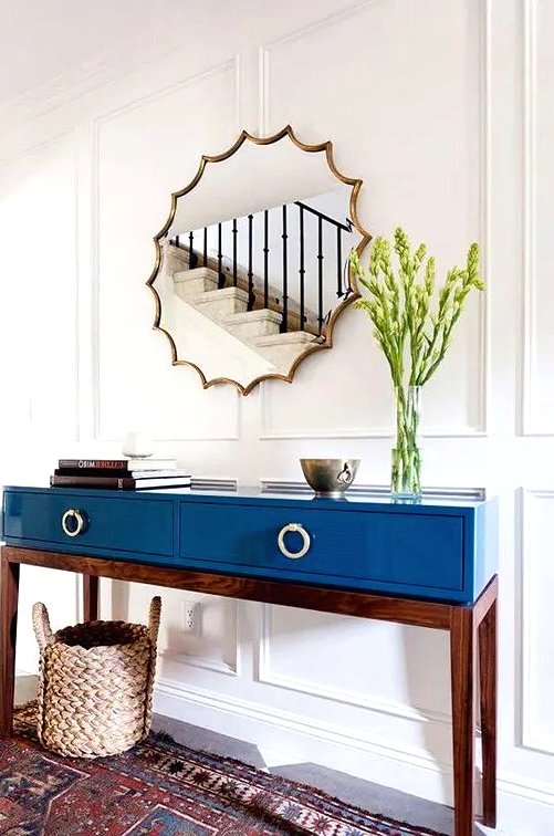 a mid-century modern table in blue lacquer finish with drawers, greenery in a clear vase, a bowl, a stack of books and a mirror of a very catchy shape