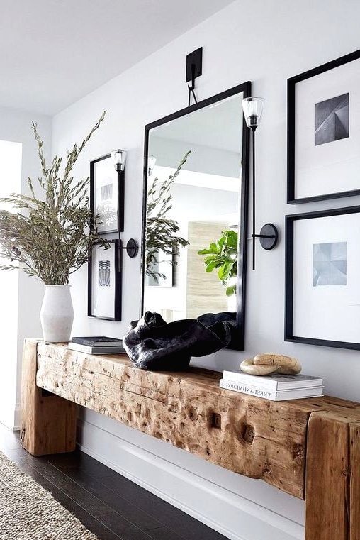 a refined contemporary space with a wood slab console table, lots of greenery branches in a vase, a black artwork, books and an oversized mirror