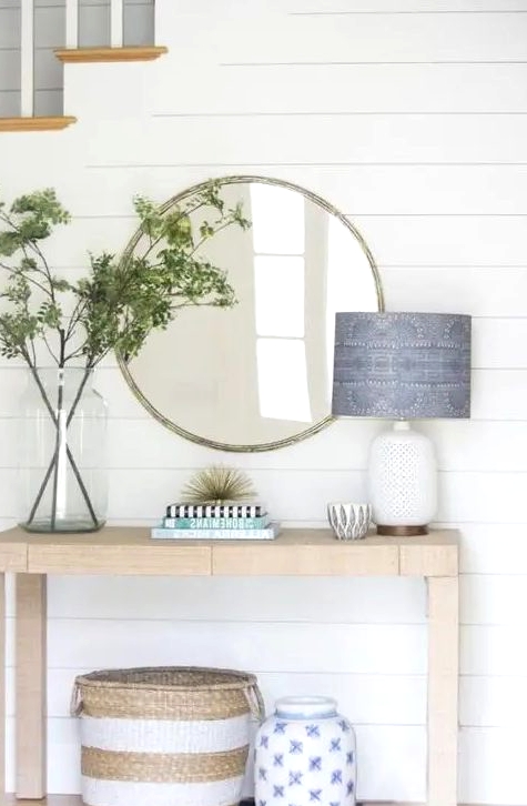 a stylish farmhouse light-stained console table, a round mirror, greenery branches in a large jar, a lamp with a blue printed lampshade and a basket under the table