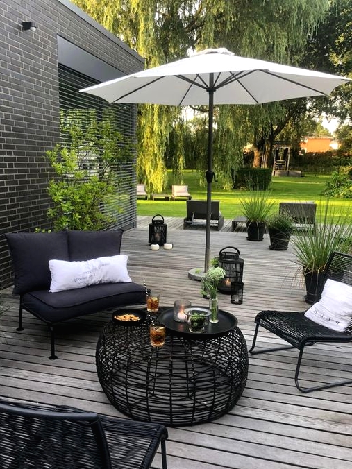 a contrasting terrace with a grey wooden deck, black metal furniture, white pillows, an umbrella and a black wicker table