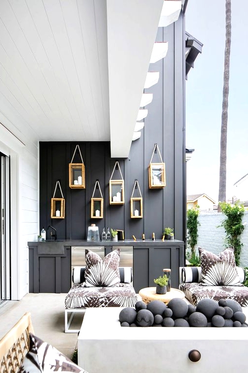 a stylish contemporary outdoor space with a black wall and a bar counter, white chairs with printed upholstery, a white fireplace wiht black pebbles