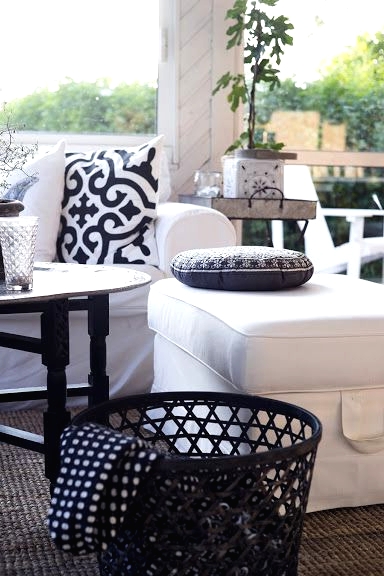 a porch with a white sectional, printed pillows, a black table and a black basket with blankets, greenery around