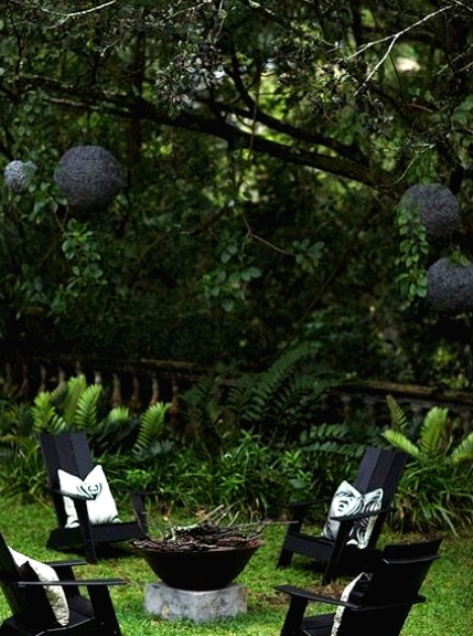 a beautiful and all-green outdoor space with a fire bowl, black chairs, printed pillows and black pendant lamps