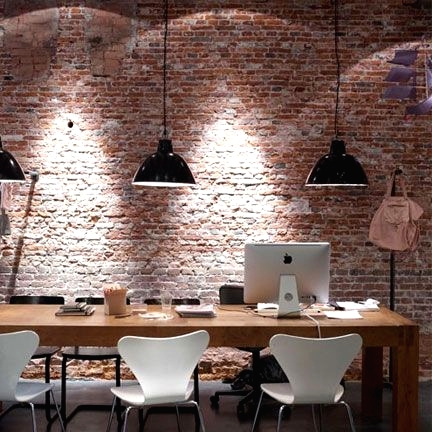 an industrial office wiht red brick wall, a large shared desk, white chairs and black metal pendant lamps