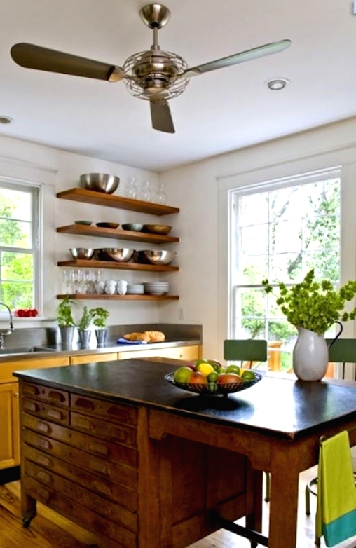 a yellow kitchen with dark countertops, open shelves instead of upper cabinets, a vintage desk as a kitchen island and green stools