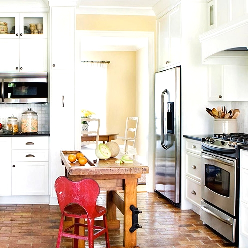a white farmhouse kitchen with a brick floor, a small stained wood kitchen island, a red chair and black countertops for a touch of contrast here