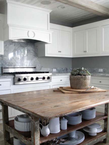 a vintage farmhouse white kitchen with a neutral stone backsplash and countertops, a large stained kitchen island that can double as a table