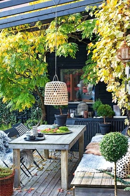 a relaxed Scandinavian deck with a built-in bench and a table, a pendant lamp, potted greenery and some trees