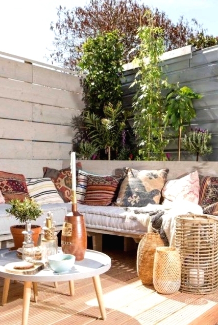 an inviting deck with a corner pallet bench and printed pillows, round tables, candle lanterns and potted greenery and climbing plants