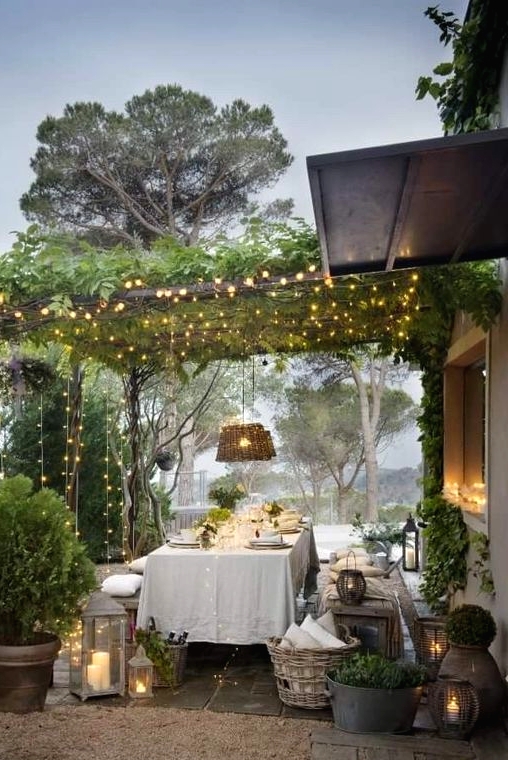 a lovely Provence outdoor dining space with elegant dining furniture, white linens, woven pendant lamps, greenery and lots of lights