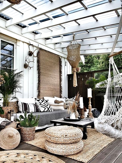 a boho outdoor living room with a glass roof, a large sofa, jute rugs and poufs, woven chairs and printed pillows