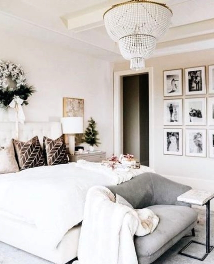 an elegant neutral bedroom with a creamy upholstered bed, a grey loveseat, a refined gallery wall, a beaded chandelier and a wreath