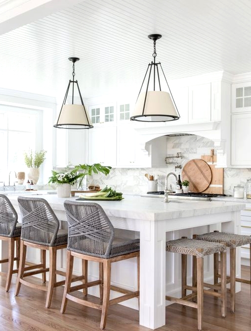 a beautiful modern kitchen with white shaker style cabinets, a large kitchen island with woven chairs and stools, pendant lamps