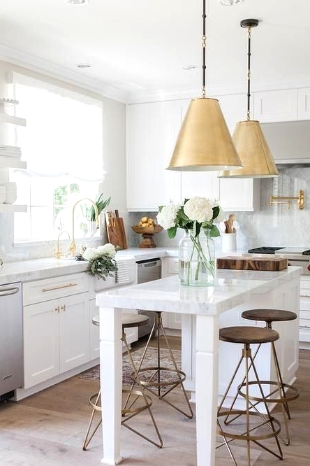 a modern and airy kitchen with white shaker style cabinets, a small kitchen island that doubles as a table, brass pendant lamps