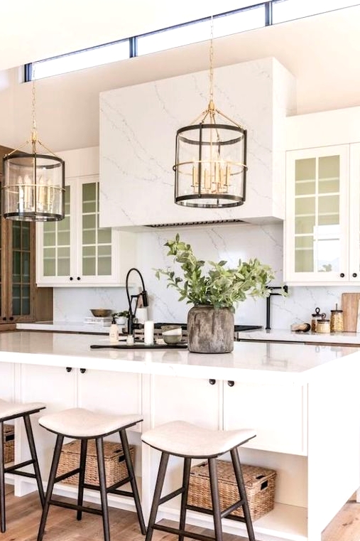 a pretty modern farmhouse kitchen done in creamy shades, with a white marble backsplash and a ahood, a large kitchen island and neutral stools