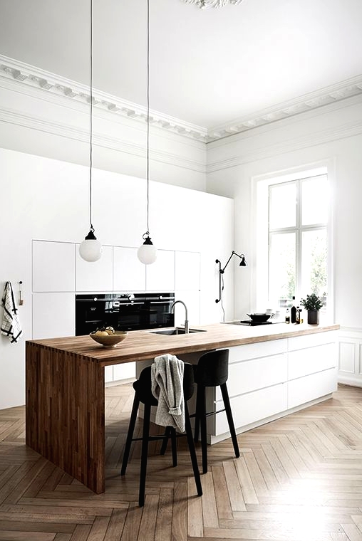 a refined minimalist kitchen with sleek white cabinets, a large kitchen island with a wooden waterfall countertop, black stools and pendant lamps