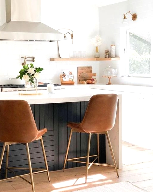 a small and pretty modern kitchen with white lower cabinets, open shelves, a black planked kitchen island and leather and brass stools