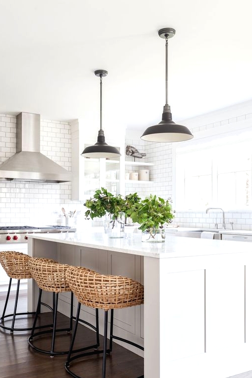 an airy white kitchen with a large kitchen island, black metal pendant lamps, woven stools on black metal bases and greenery