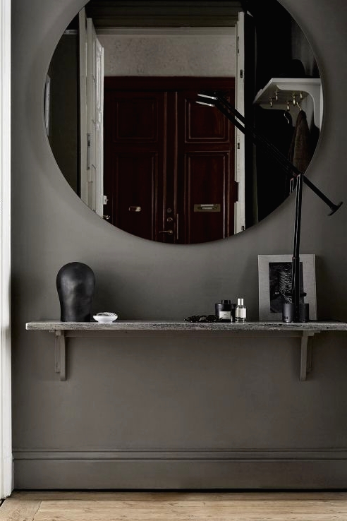 a slate grey entryway with a round mirror, a shelf, a table lamp and some art is a very chic and minimal space