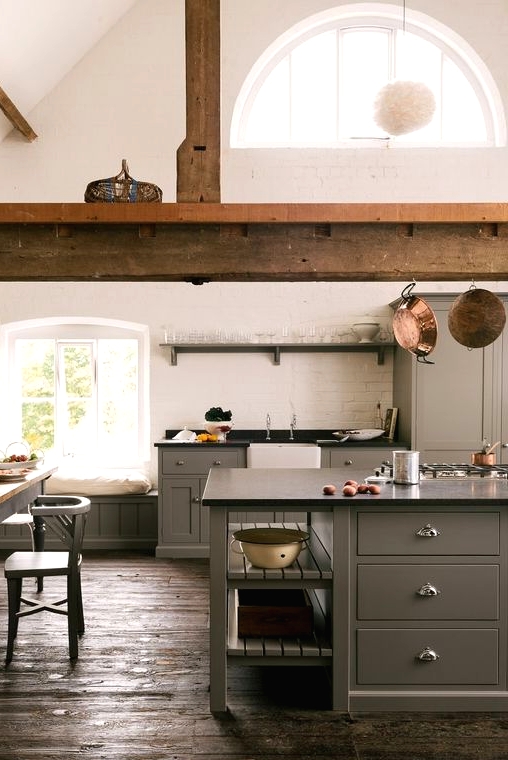 a barn kitchen with grey cabinets, a large wooden beam, black countertops, a vintage dining set and white brick walls