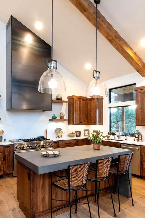a modern barn kitchen with stained cabinets, white stone and concrete countertops, a shiny hood, wooden beams and pendant lamps
