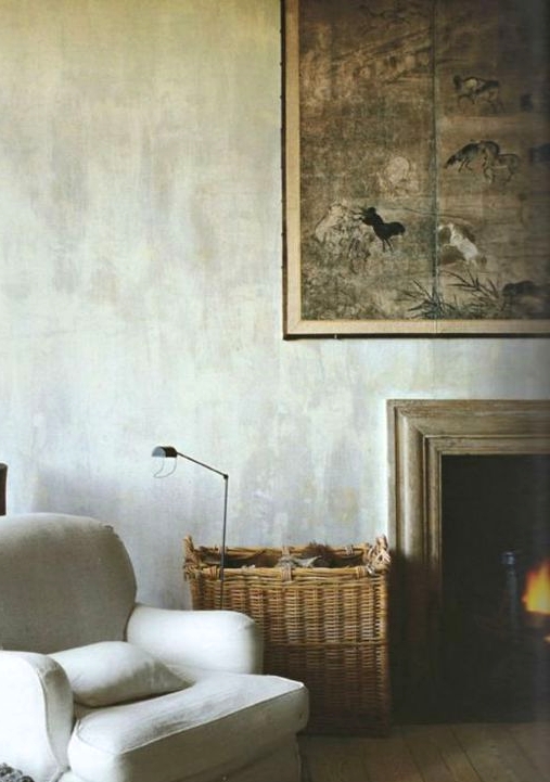 a cozy space with grey limewashed walls, a fireplace, a creamy chair, a basket with firewood and an artwork