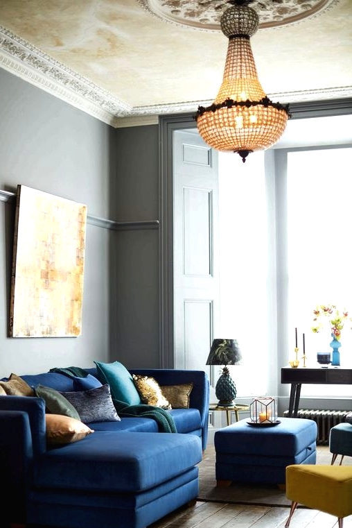 a beautiful and refined living room with grey walls, a navy sectional, a navy pouf and yellow and blue stools, a crystal chandelier and a statement artwork