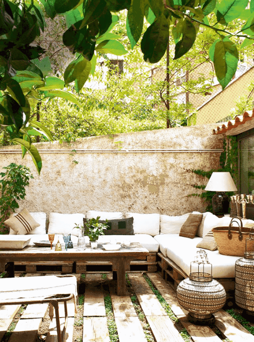 a relaxed outdoor Mediterranean space with a grass and wood plank floor, a pallet corner sofa with neutral upholstery, a wooden table and a daybed, some Moroccan lanterns