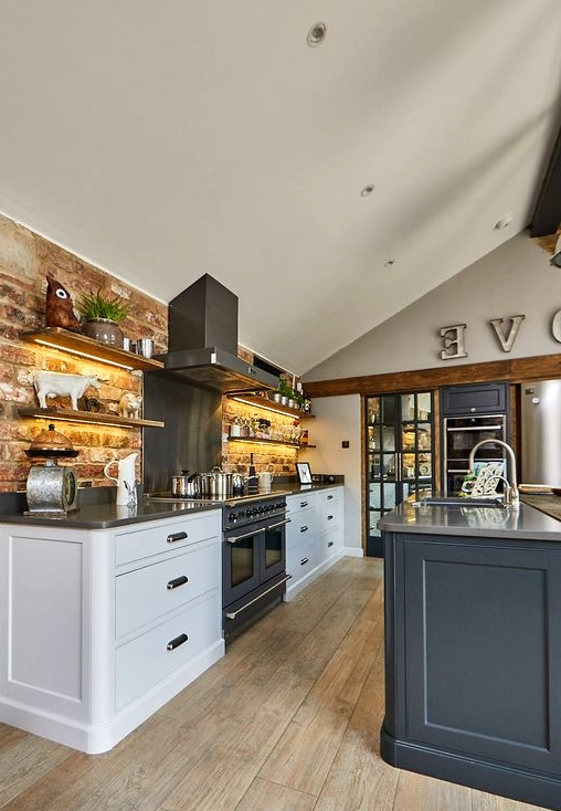 a barn kitchen with a brick accent wall, white cabinets and a stone blue kitchen island, wooden beams and stainless steel appliances