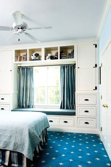 a blue and white bedroom with blue walls and a navy rug, a bed with blue bedding, a neutral storage unit with drawers and blue curtains and an upholstered bench