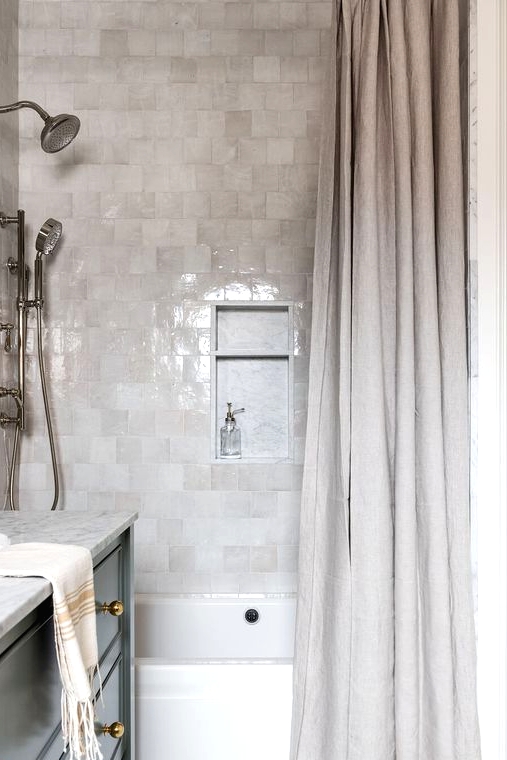 a chic contemporary bathroom clad with light grey and neutral zellige tiles, with a neutral shower curtain and a black vanity
