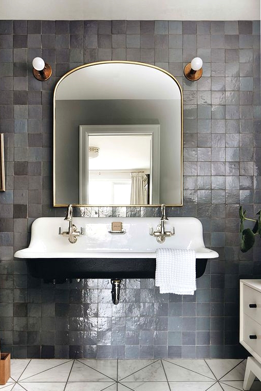 a vintage bathroom with grey zellige tiles, a black floating sink, a curved mirror, brass and gold fixtures and marble tile on the floor